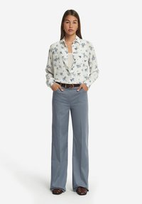 Vit långärmad blus med blått blommigt mönster, ihopparad med ljusgrå vida byxor och brunt bälte. Modellen står med händerna i fickorna.