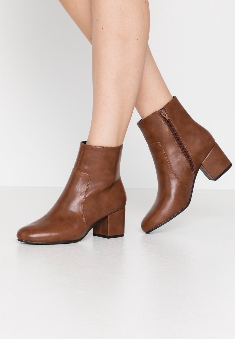 Bottes chevilles en cuir marron avec talons bloc portées sur des jambes croisées, avec un détail de fermeture éclair latérale sur un fond de sol clair.