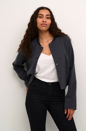 Veste bomber gris foncé avec des boutons-pression, col rond et manches longues, associée à un haut blanc et un jean noir. Tissu texturé uni.