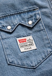 Lys blå denimstoff som viser en lomme med sølvknapper. Merket har påskriften "Levi's AUTHENTIC WESTERN WEAR" med en tegneserie-logo og "PIXAR."