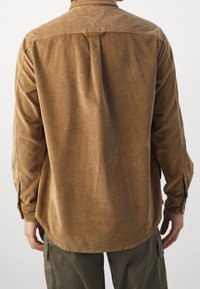 Brun kordfløjlsjakke med knaplukning på kraven, lange ærmer og et lige snit. Har markante lodrette ribber og en rundet kant.