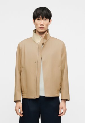 Joven de pie, con una chaqueta beige con cremallera y cuello alto, sobre una camisa blanca y pantalones oscuros, contra un fondo liso.