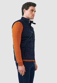 Marineblaues isoliertes Weste mit Reißverschluss, Seitentaschen und hohem Kragen über einem orangefarbenen Langarmshirt, getragen mit lässigen dunklen Jeans.