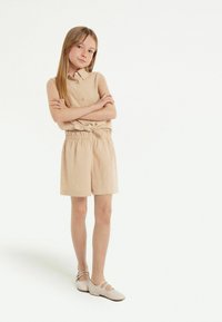 Tutina beige senza maniche con colletto, apertura con bottoni e vita elasticizzata. Abbinata a scarpe piatte con cinturini di un beige chiaro. Tessuto liscio.