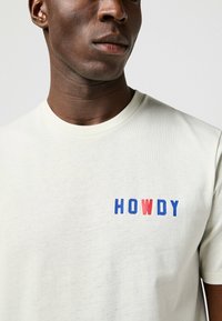 Camiseta de algodón crema clara con la palabra "HOWDY" en letras de bloque rojas y azules en el lado izquierdo del pecho. Corte casual.