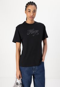 Black cotton t-shirt featuring a rhinestone-embellished "Hugo" logo; relaxed fit with short sleeves and a crew neckline. Paired with denim jeans.