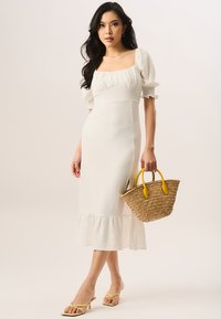White midi dress with puff sleeves, ruched bodice, and ruffled hem. Woven straw handbag with yellow handles. Minimalist footwear.