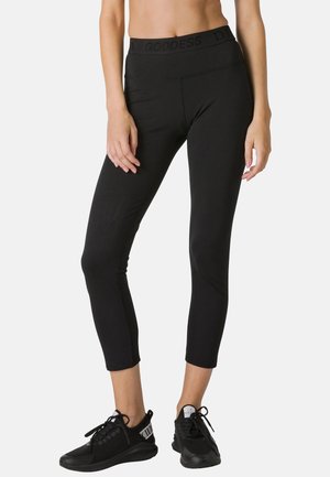 Woman wearing black cropped leggings with "GODDESS" waistband detail and black sneakers, standing against a plain light background.