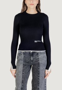 Haut noir à manches longues côtelé avec des accents de poignet blancs, arborant le logo "Karl Lagerfeld Jeans" à l'avant, associé à un pantalon en denim gris.