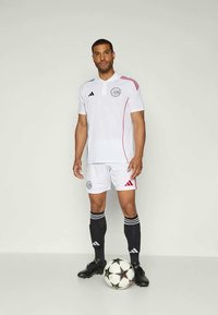 Uniforme da calcio bianco Adidas con polo a maniche corte con dettagli rossi e logo, abbinato a pantaloni corti e calzettoni neri con strisce bianche. Una palla da calcio è posata nelle vicinanze.