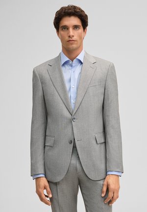 Mann trägt einen hellgrauen, maßgeschneiderten Anzug mit einem hellblauen Hemd und steht vor einem einfarbigen grauen Hintergrund.