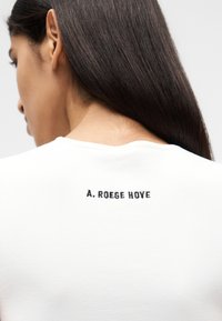 Γυναίκα με μακριά ίσια σκουρόχρωμα μαλλιά, ντυμένη με λευκό πουκάμισο που έχει τυπωμένο το κείμενο "A. ROEGE HOVE" σε μαύρο χρώμα στην άνω πλάτη.