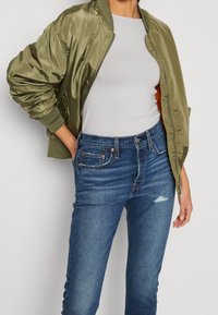 Chaqueta bomber de satén verde oliva con puños acanalados, llevada sobre una camiseta gris ajustada y jeans azules desgastados, con bolsillos frontales.