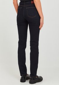 Slim-fit dark blue denim jeans with a mid-rise waist, rear pockets, and contrasting orange stitching, paired with black shoes.