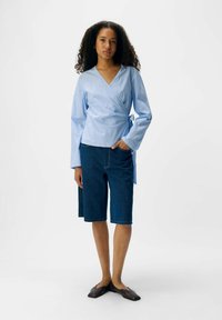 Top avvolgente a righe azzurro chiaro con maniche lunghe, abbinato a pantaloni corti in denim scuro fino al ginocchio e scarpe slip-on scure. Design semplice.