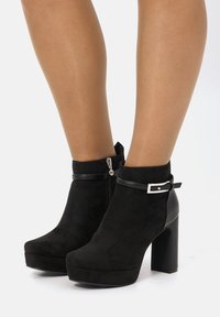 Stivaletti neri in suede con tacco alto a blocco, chiusura con zip, dettagli in pelle e una fibbia sulla cinghia alla caviglia.