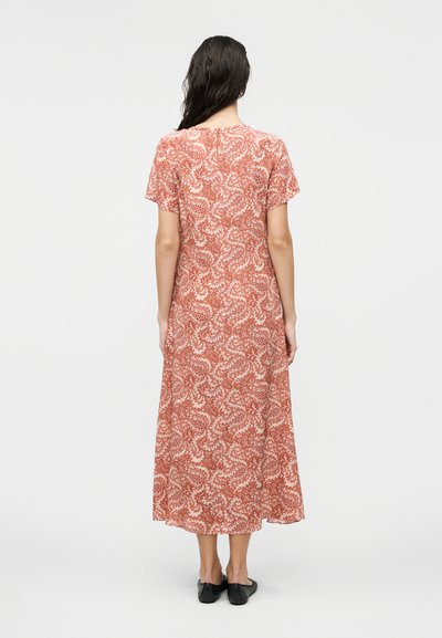 Γυναίκα με μακριά σκούρα μαλλιά στέκεται με την πλάτη, φορά φόρεμα μήκους μέχρι τη γάμπα με κοντά μανίκια σε σκουριά απόχρωση με λευκό φλοράλ μοτίβο παϊέτας και μαύρα φλατ παπούτσια.