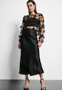 Zwart tweedelig outfit bestaande uit een doorzichtige top met lange mouwen, versierd met bloemenapplicaties, en een satijnen midi-rok. Sleehak sandalen maken de look compleet.