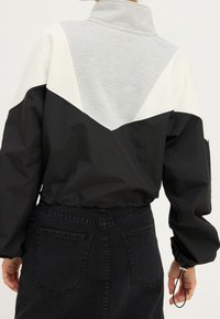 Korte jas met hoge kraag. Kleurvlakken: zwart, lichtgrijs en crèmekleurig. Gestructureerde stof, elastische zoom en geplooide mouwen met koord.