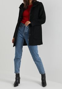 Svart ullblandad kappa med huva, framtida snap-stängning och stora sidofickor, lagd över en röd ribbad topp och blå jeans. Svarta ankelstövlar.