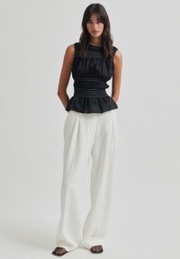 Juoda be rankovių peplumo tipo palaidinė su susiraukusiu audiniu ir baltais siuvinėjimo akcentais, suderinta su laisvomis baltomis kelnėmis ir juodais tinkliniais loaferiais.