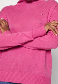 Roze gebreide trui met een hoge halslijn, geribbelde boorden en zoom. Heeft een klein logo op de borst. Zachte textuur en losse pasvorm.
