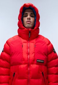 Chaqueta de plumas roja con cuello alto y cierre de cremallera, que presenta un interior forrado en azul, parches de acento negros y cuadrados de aislamiento.