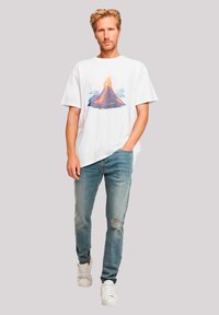Weißes Baumwoll-T-Shirt mit einem bunten Vulkan-Graphic, kombiniert mit hellblauen, ausgebleichten Jeans und weißen Sneakers.