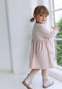 Svetloružové šaty s dlhými rukávmi, naškrobený pás, kvetinová výšivka, doplnené béžovými pletenými pančuchami a striebornými topánkami. Jasné okno v pozadí.