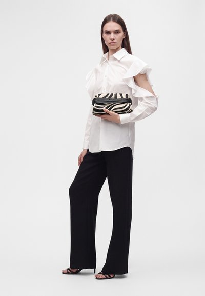 Femme portant un chemisier blanc à épaules volantées avec des découpes, pantalon large noir, sandales noires, tenant une petite pochette imprimée zèbre.