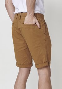 Homme portant un short marron retroussé et un t-shirt blanc, la main droite placée dans la poche arrière du short.