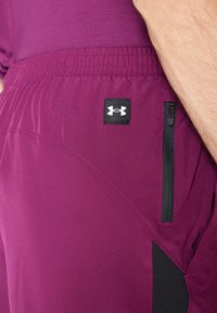 Burgundi sportnadrág elasztikus derékkal, oldalsó cipzáras zsebbel és fekete hálós díszítéssel. Az Under Armour logó a bal oldalon.