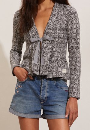 Black and white patterned jacket with a peplum design and gray tie at the waist, paired with rolled denim shorts featuring star embroidery.