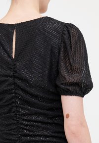 Haut noir texturé avec des manches bouffantes et une ouverture en keyhole à l'arrière. Le tissu présente une surface brillante et à motifs avec un détail de fronces.