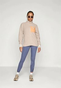 Light beige zip-up jacket with an orange front pocket, paired with fitted blue leggings and beige sneakers, featuring a smooth texture.