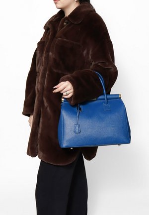 Borsa a mano in pelle blu con finitura testurizzata, forma strutturata, manico superiore e dettagli dorati. Abbinata a un cappotto in pelliccia sintetica marrone.
