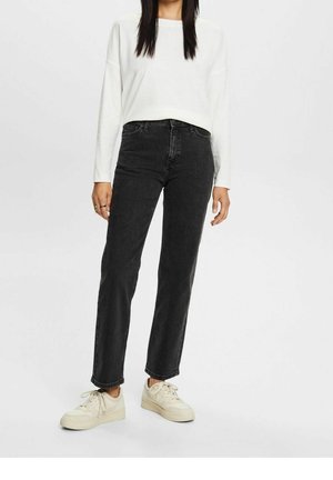 Zwarte spijkerbroek van denim met een rechte pasvorm, hoge taille en minimale details, gecombineerd met een losvallend wit langarmtop en crèmekleurige sneakers.