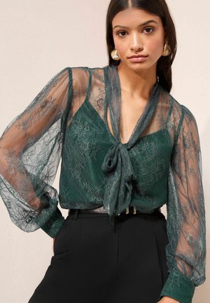 Grünspitze Bluse mit transparenten langen Ärmeln und Bindeband am Ausschnitt. Verfügt über aufwendige Blumenmuster und eine figurbetonte Passform.