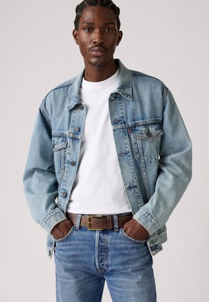 Light blue denim jacket with buttoned front, two chest pockets, and cuffed sleeves, worn over a white t-shirt and blue jeans, complemented by a brown belt.