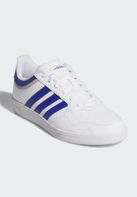 Zapatilla deportiva blanca con acentos en azul y plata, que presenta tres rayas azules, un sistema de cordones en la parte delantera, una caja de dedos perforada y una suela de goma texturizada.