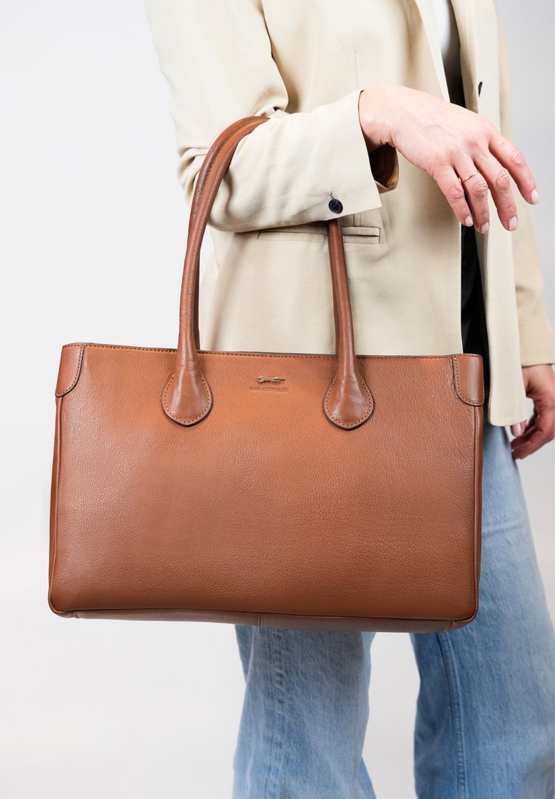 Sac fourre-tout en cuir marron avec une texture lisse, une forme allongée et des côtés structurés. Dispose de deux poignées et d'un logo subtil à l'avant.