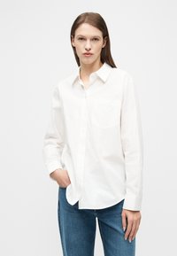 Γυναίκα με ίσια καστανά μαλλιά, φοράει λευκό, άνετο πουκάμισο με κουμπιά και μπλε τζιν, στέκεται με το ένα χέρι στην τσέπη σε λευκό φόντο.