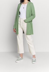 Cardigan verde a maglia sopra una t-shirt bianca, abbinato a pantaloni culotte crema e sneakers verde chiaro, con dettagli in pizzo e un fit casual.