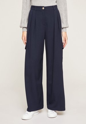 Granatowe spodnie o szerokich nogawkach z wysokim stanem, z przodu z plisami, boczne kieszenie cargo, zapięcie na złoty guzik, noszone z białymi trampkami i szarym swetrem z warkoczowym splotem.