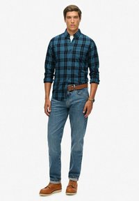 Ruitenoverhemd met knopen in donkerblauw en zwart, blauwe jeans, leren bruine riem en bruine schoenen. Model staat rechtop tegen een effen achtergrond.