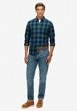 Camisa de cuadros de botón en azul oscuro y negro, jeans azules, cinturón de cuero marrón y zapatos marrones. El modelo está de pie contra un fondo liso.