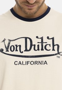 T-shirt à manches courtes crème avec un col en ribbed bleu marine. Présente un grand logo noir imprimé "Von Dutch CALIFORNIA" dans un style vintage.
