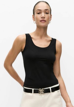 Woman wearing a black sleeveless top, white pants, and a black belt with a decorative gold buckle, standing against a plain white background.