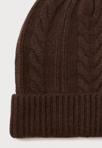 Berretto di lana lavorato a maglia marrone con motivi a treccia e orlo a costine ripiegato su uno sfondo bianco.