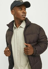 Chaqueta de plumas marrón oscuro con un alto cuello sobre una camisa de cuadros beige. El modelo lleva una gorra azul marino. Tejido suave, cierre de cremallera y bolsillos laterales.
